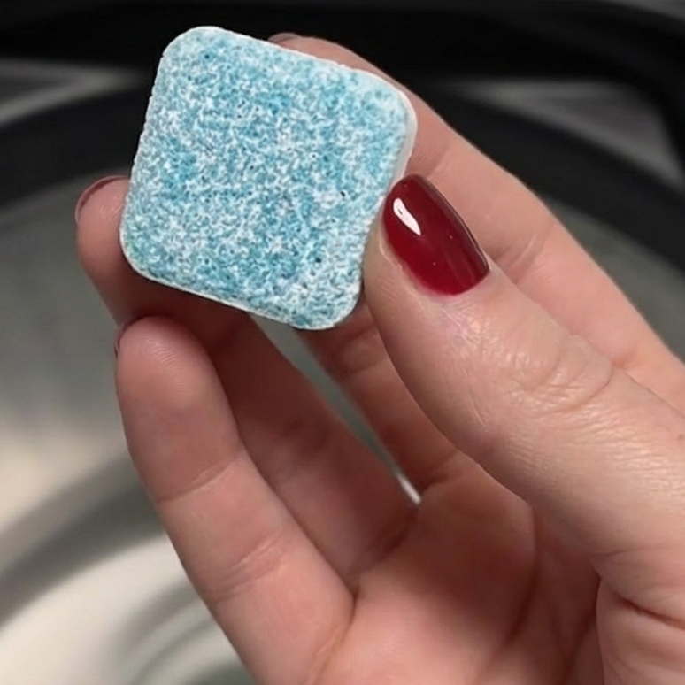 A square blue and white washing machine cleaner tablet held over a washer drum